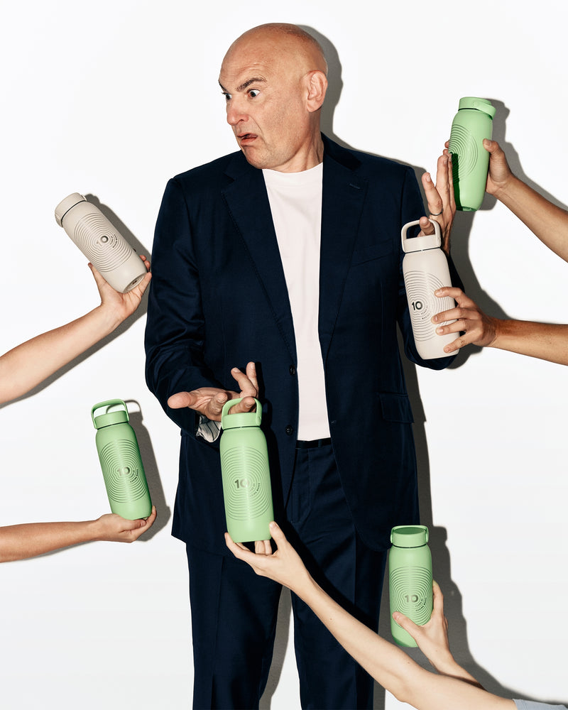 Hombre con traje oscuro rodeado por varias manos que sostienen botellas de agua verdes y blancas.
