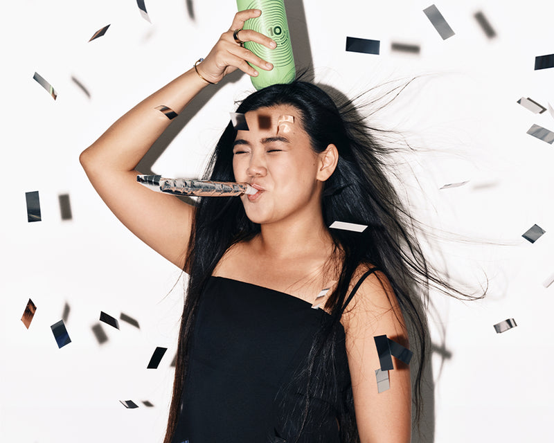 Mujer con un vestido negro celebra con un cuerno de fiesta y un spray verde, rodeada de confeti.
