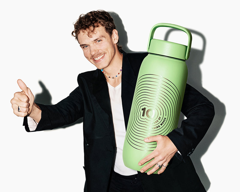 Hombre sonriente con blazer sostiene una gran botella de agua reutilizable verde con asa y da un pulgar hacia arriba.