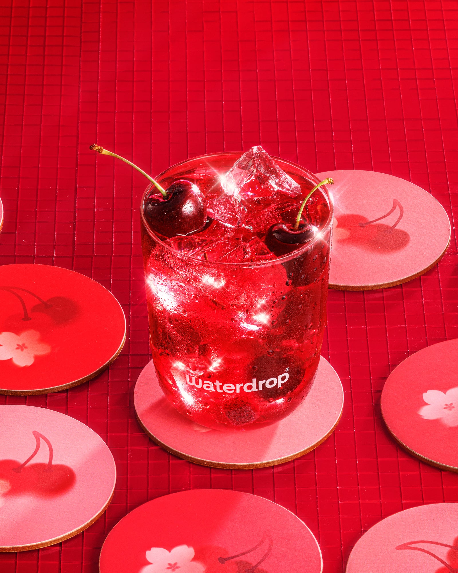Bebida roja fría con hielo y dos cerezas en un vaso sobre fondo rojo con posavasos de cerezas.