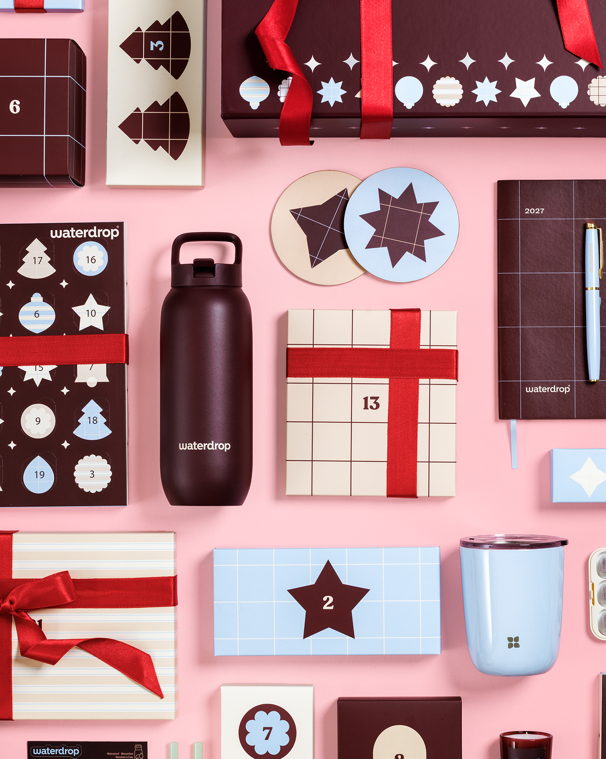 Varias cajas de regalo, un calendario de Adviento, una botella, una taza y otros regalos envueltos sobre un fondo rosa.