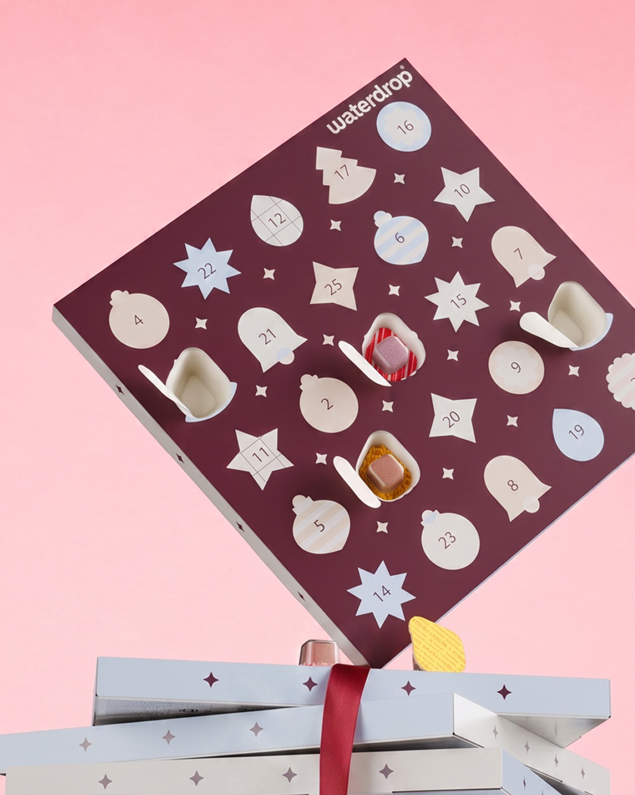 Calendario de Adviento morado con puertas numeradas en forma de estrellas sobre un fondo rosa, situado sobre dos cajas regalo.