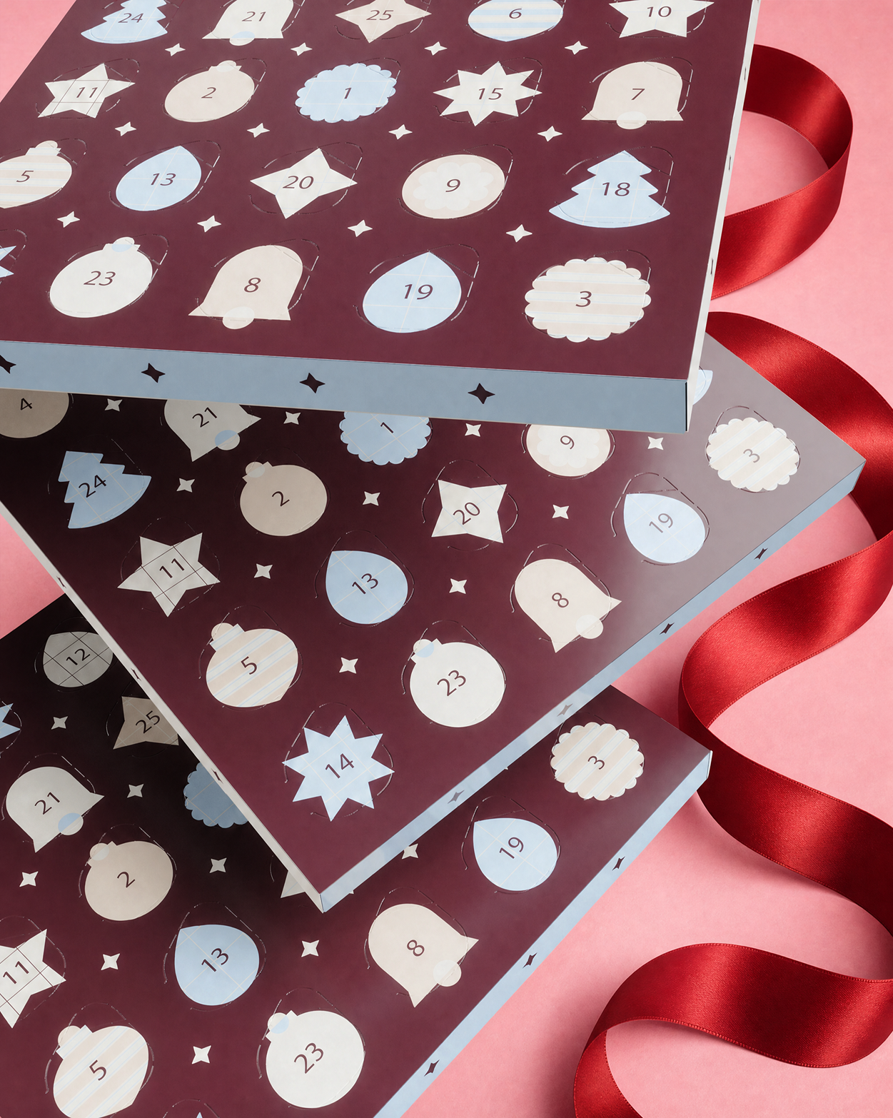 Tres cajas de calendario de Adviento apiladas con puertas numeradas y formas decorativas, atadas con una cinta roja sobre un fondo rosa.