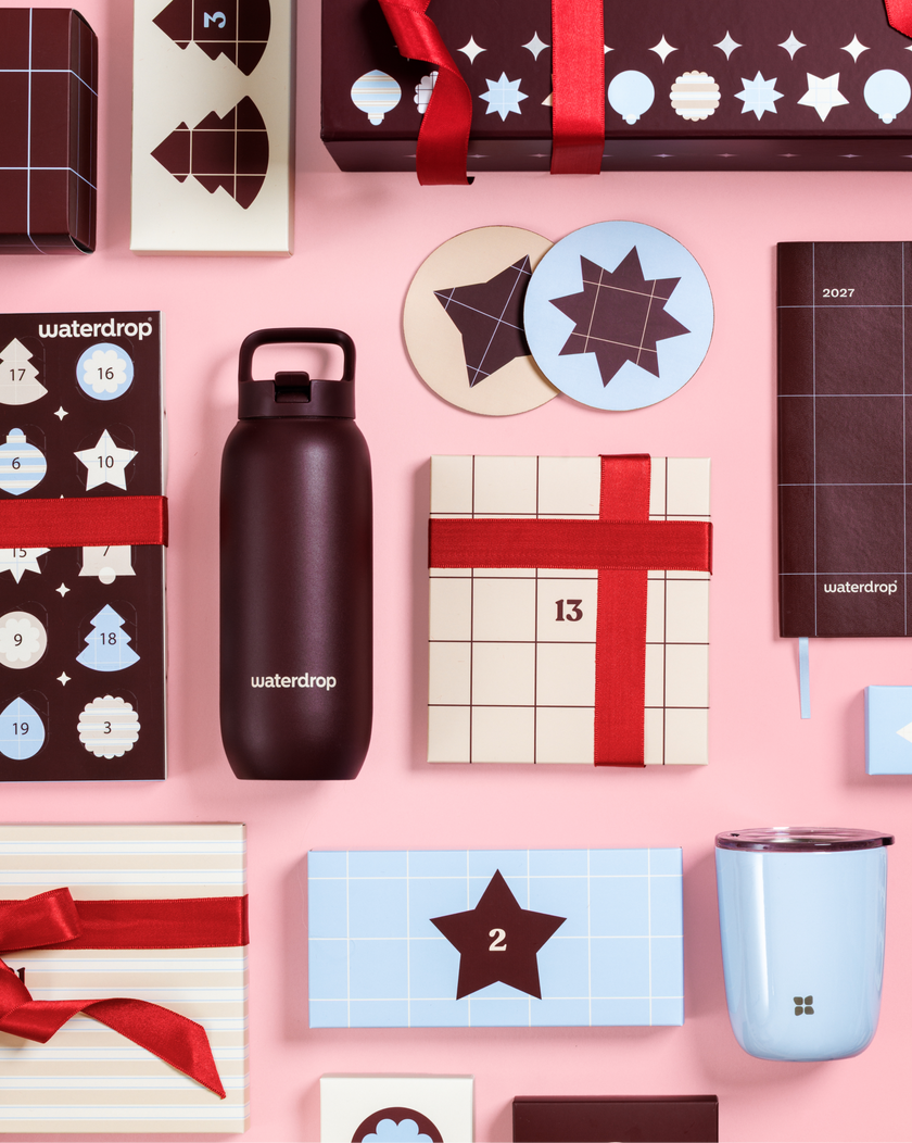 Calendarios de Adviento y cajas regalo con lazos rojos sobre un fondo rosa, junto a una botella negra y una taza azul claro.