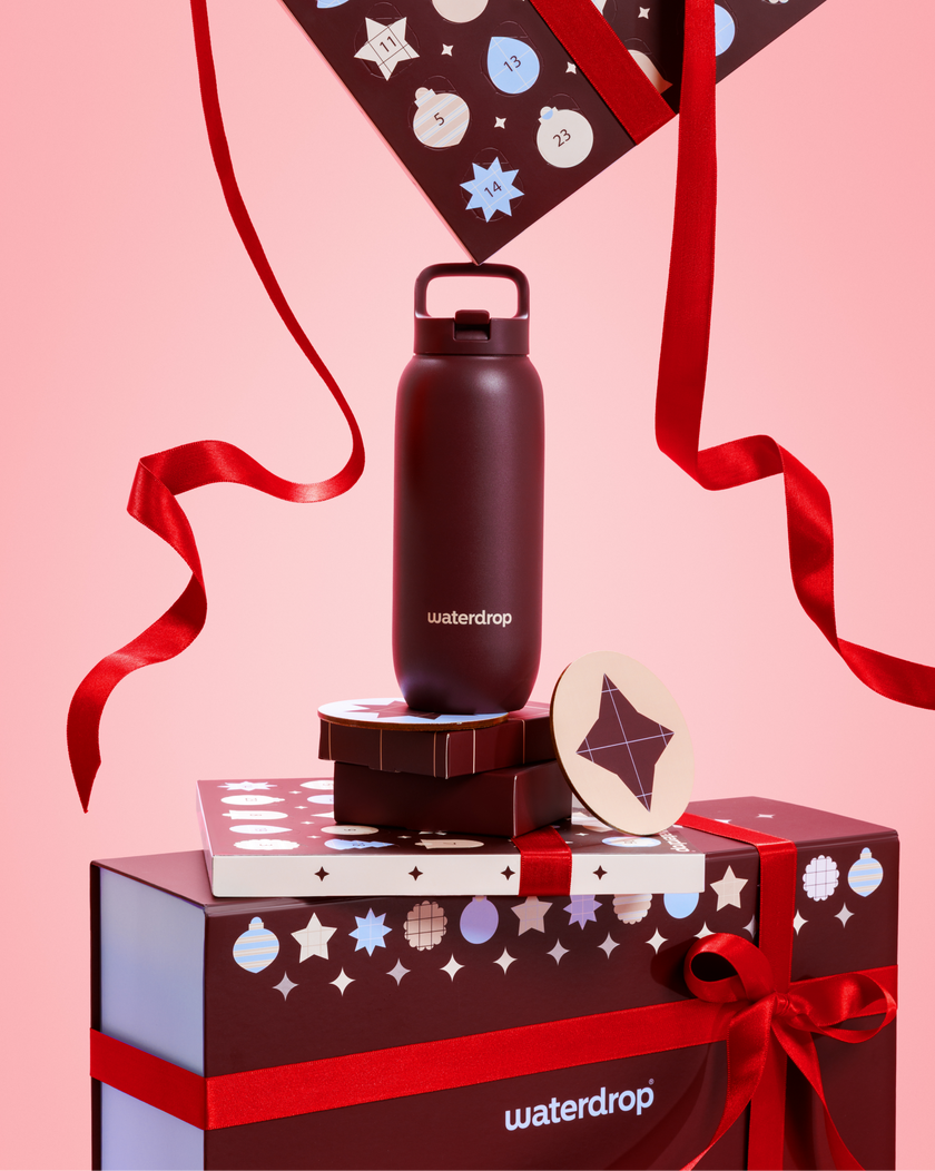 Botella de agua negra con tapa y asa, sobre cajas de regalo apiladas atadas con lazos rojos, contra un fondo rosa.