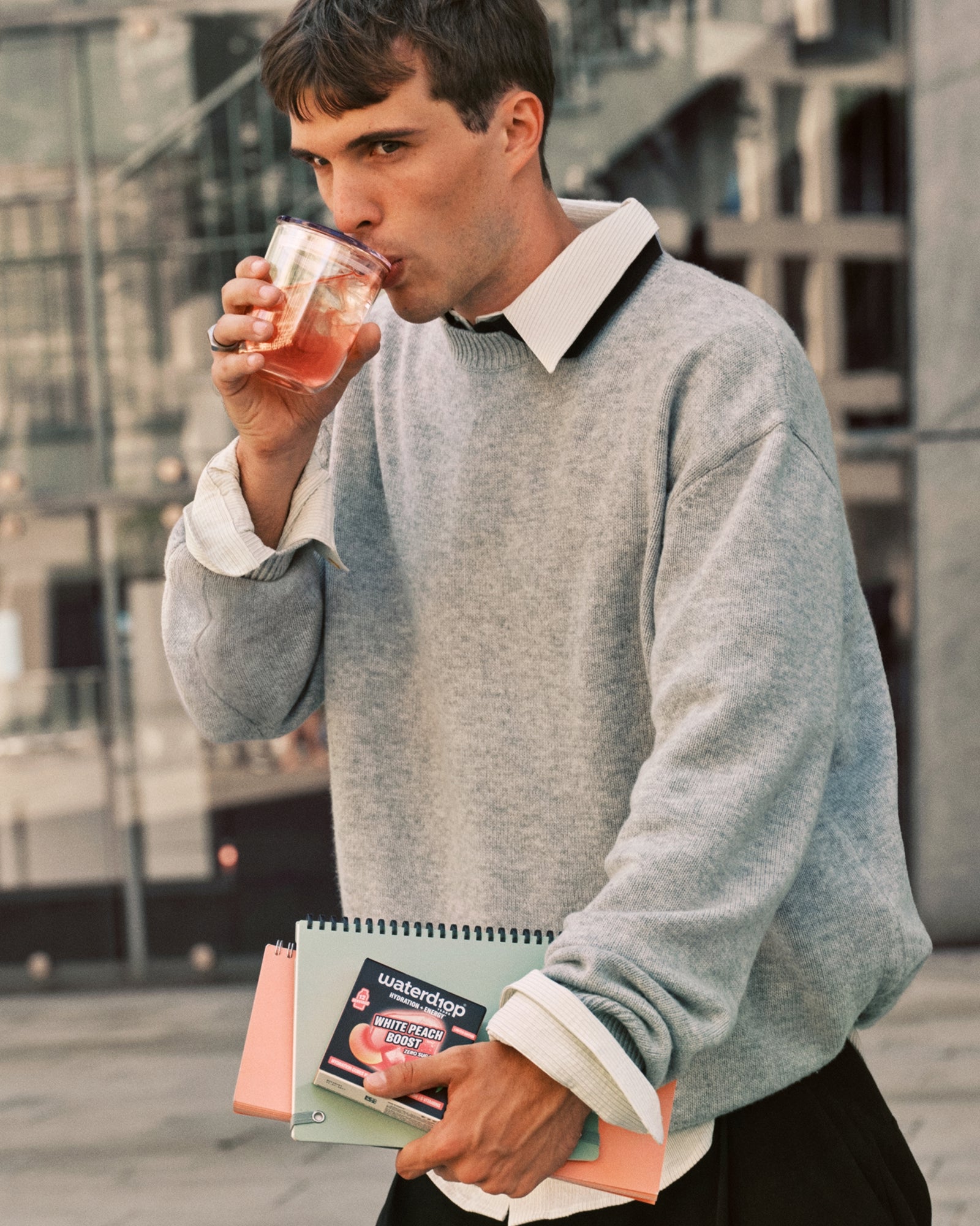 Hombre con suéter gris bebe de una taza mientras lleva cuadernos.