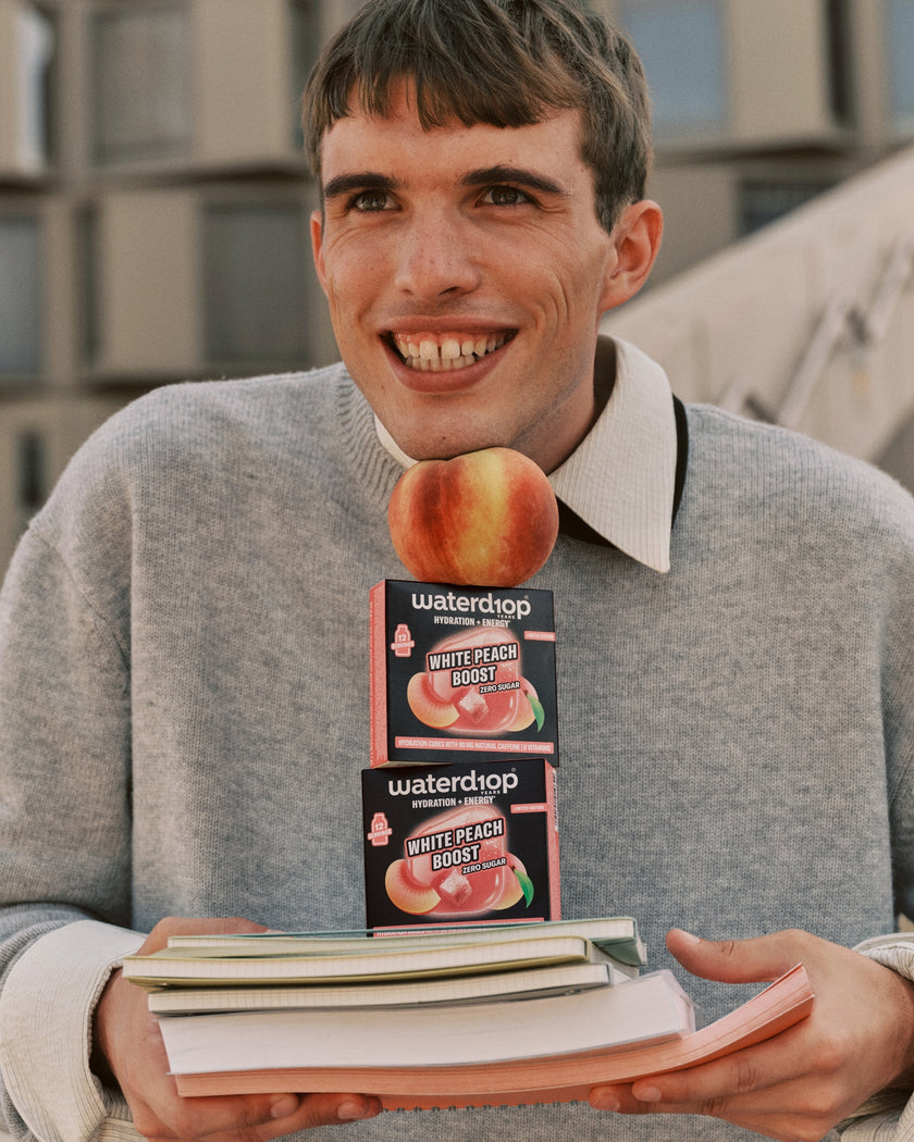 Joven con suéter gris sosteniendo dos cajas de sabor melocotón sobre una pila de libros.
