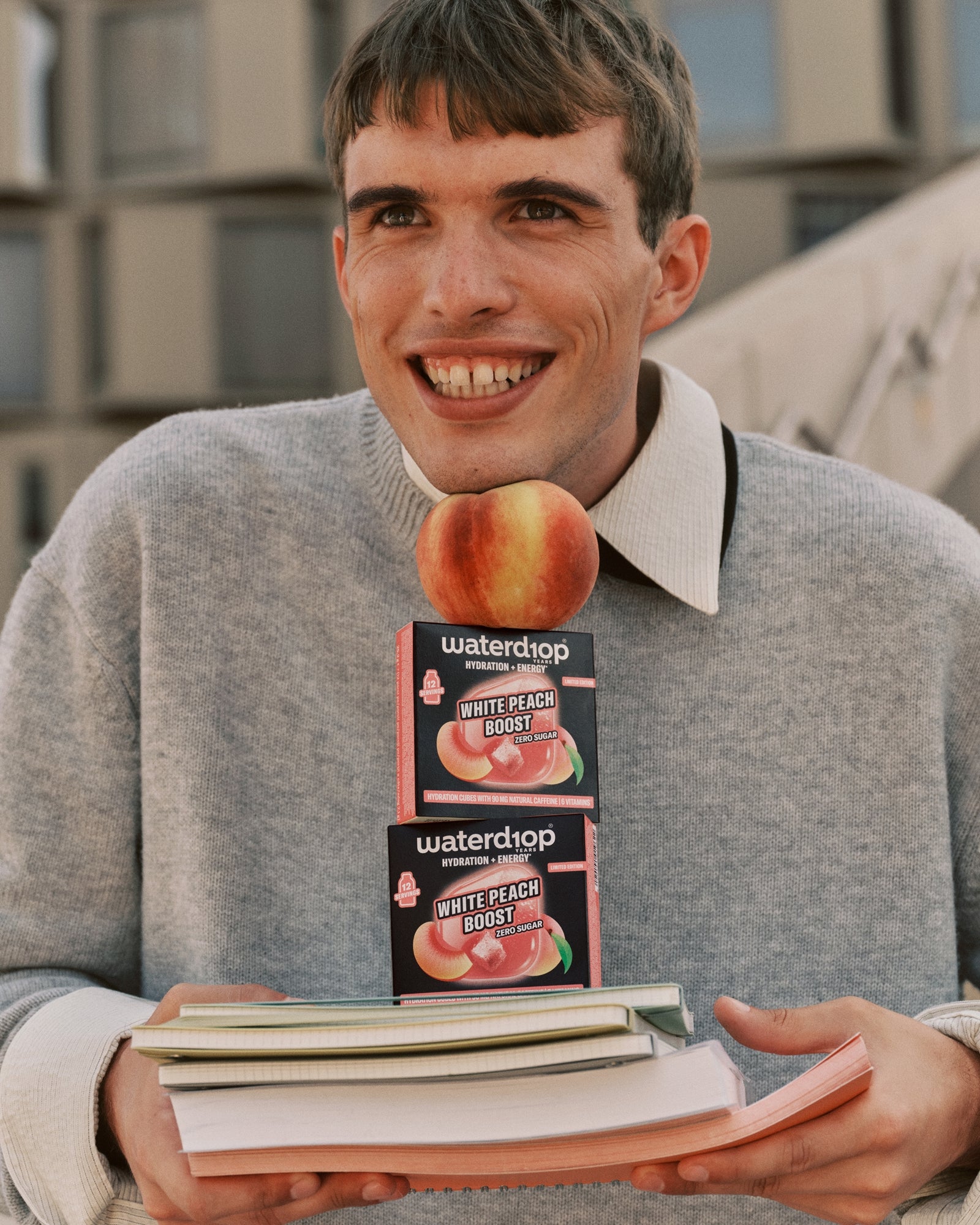 Joven con suéter gris sosteniendo dos cajas de sabor melocotón sobre una pila de libros.