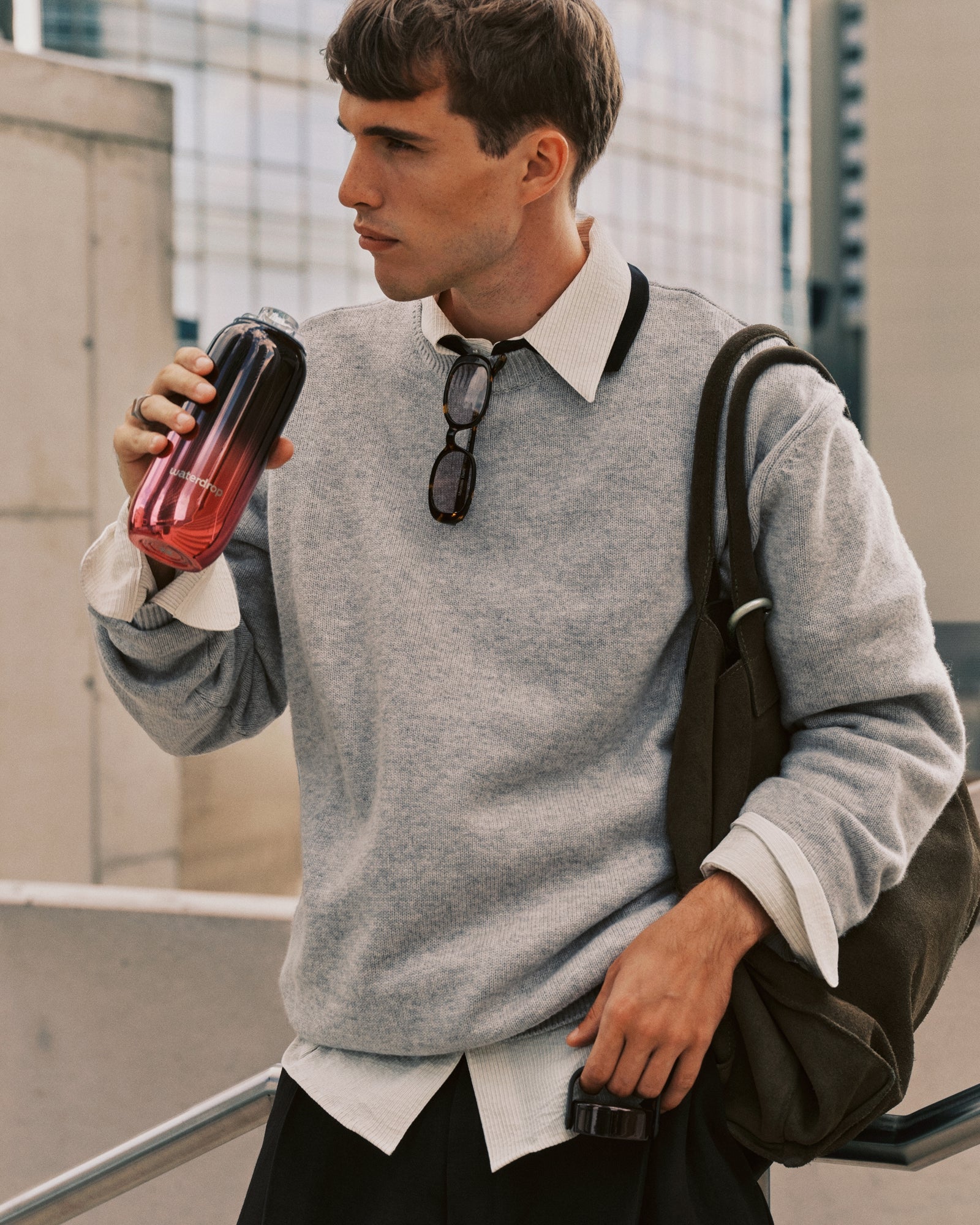 Hombre joven con suéter gris y camisa blanca lleva una mochila mientras bebe de un vaso térmico con degradado.