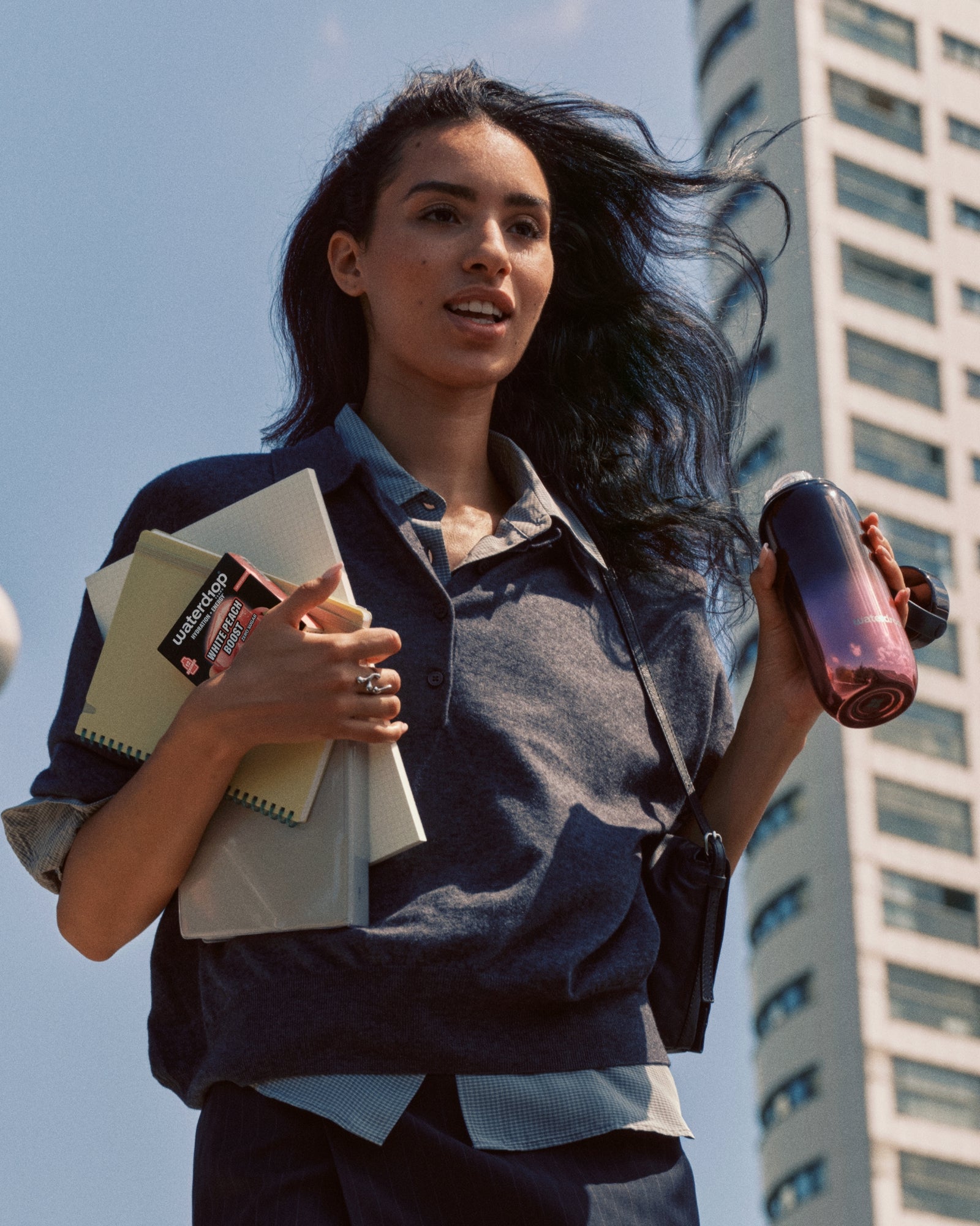 Mujer joven caminando al aire libre en una ciudad, lleva cuadernos y una carpeta, y sostiene una botella.