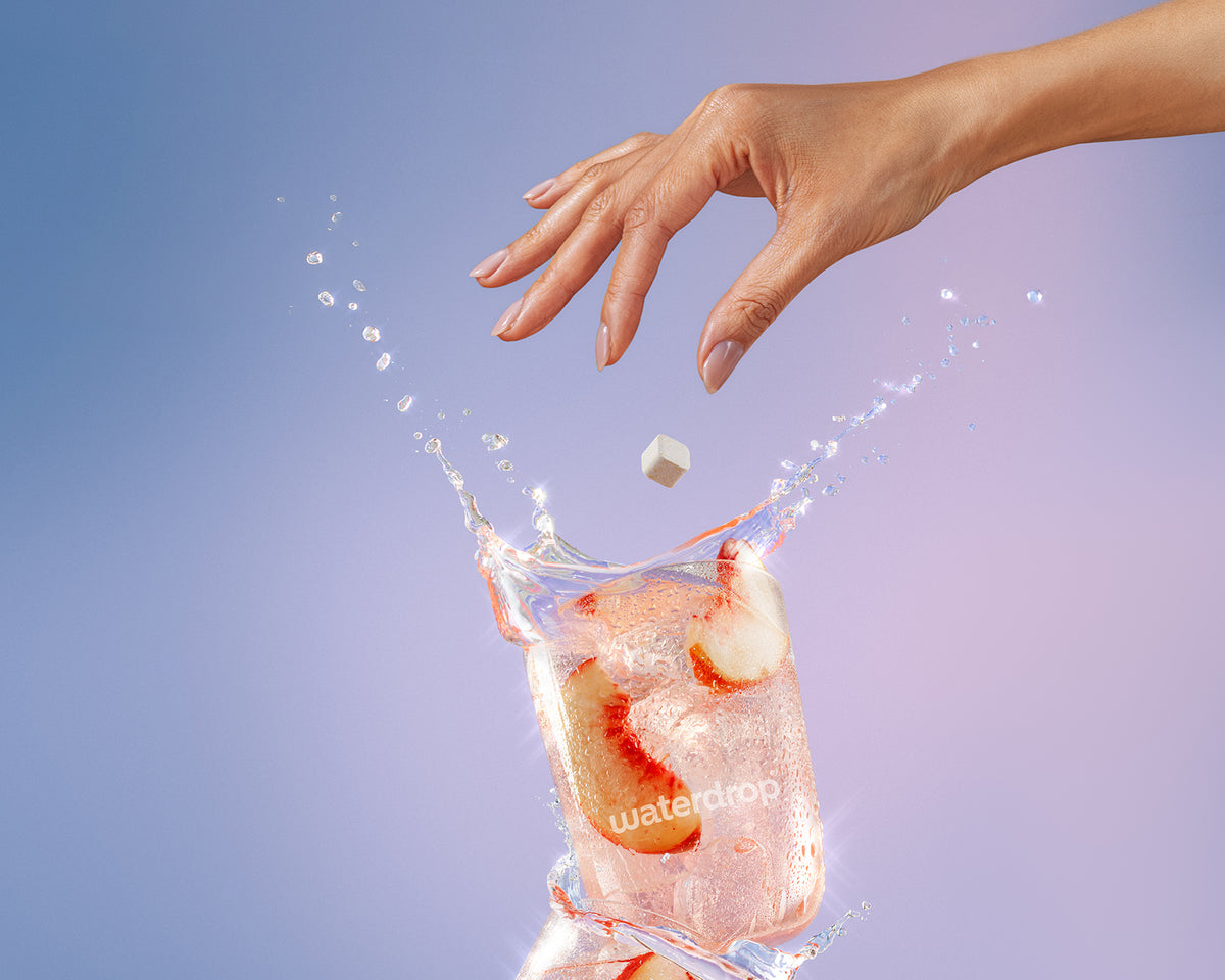 Una mano deja caer un cubo de hielo en un vaso de agua con hielo, provocando una salpicadura.