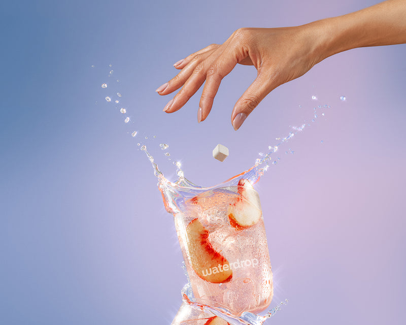 Una mano deja caer un cubo de hielo en un vaso de agua con hielo, provocando una salpicadura.