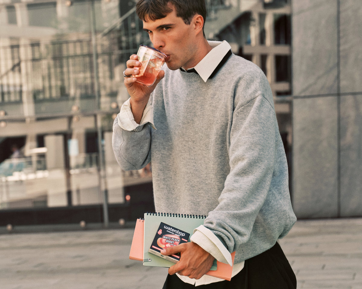 Hombre con suéter gris bebe de un vaso mientras sostiene un cuaderno al aire libre en un entorno urbano.