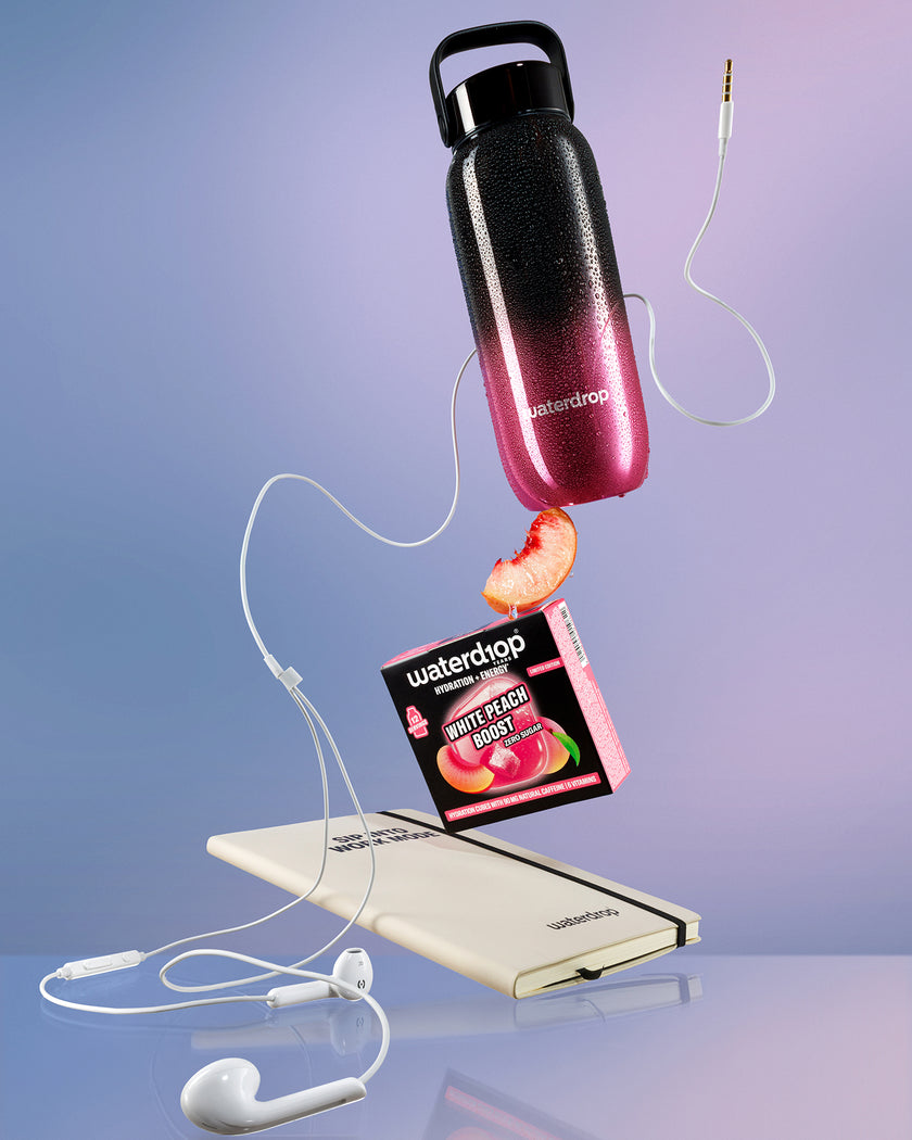 Exhibición flotante de una botella en degradado de negro a rosa sobre una batería externa blanca, conectada por cables blancos con una rodaja de durazno cerca.