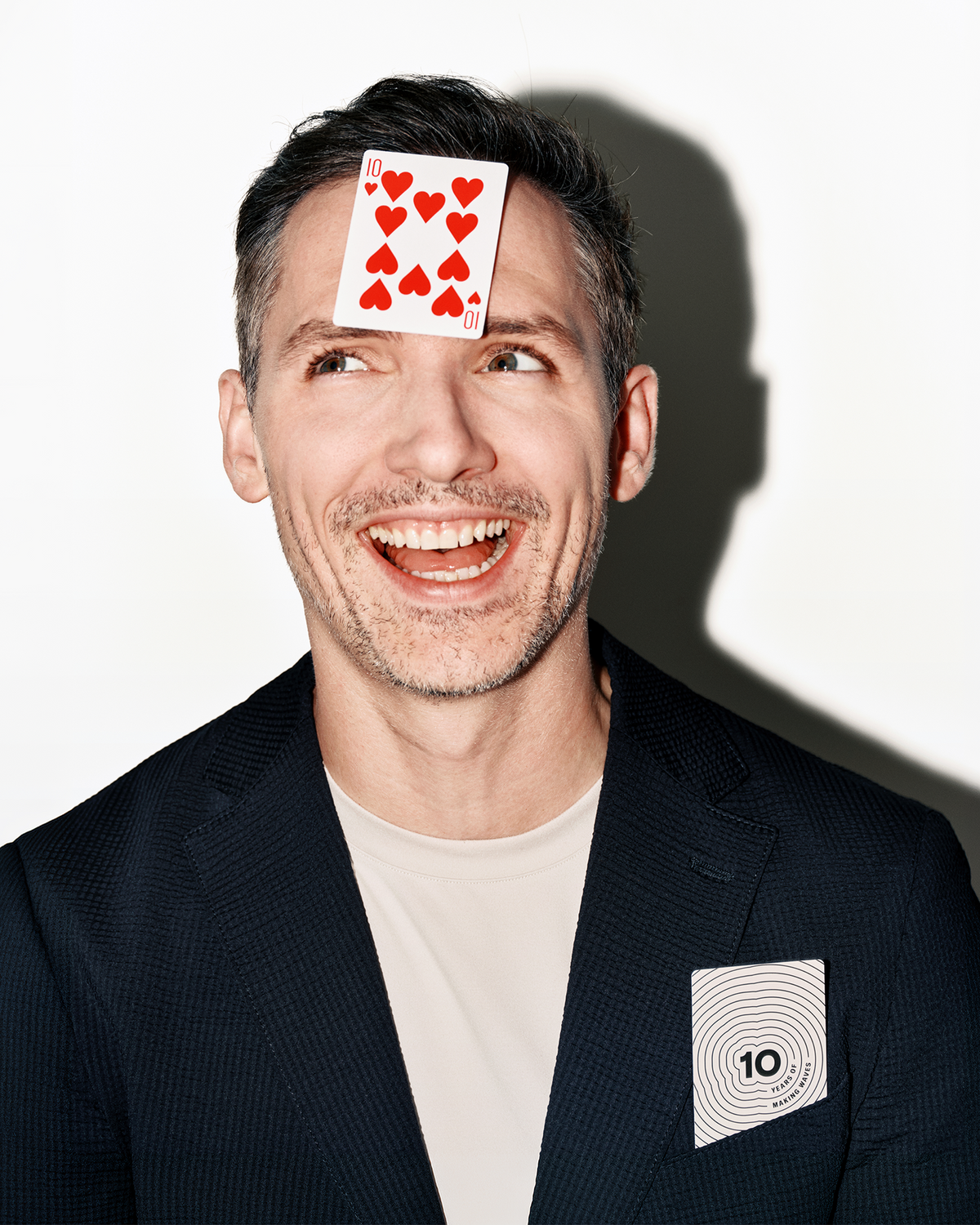 Hombre sonriendo con chaqueta oscura y camisa clara tiene una carta de juego en la frente y otra en el bolsillo del pecho.