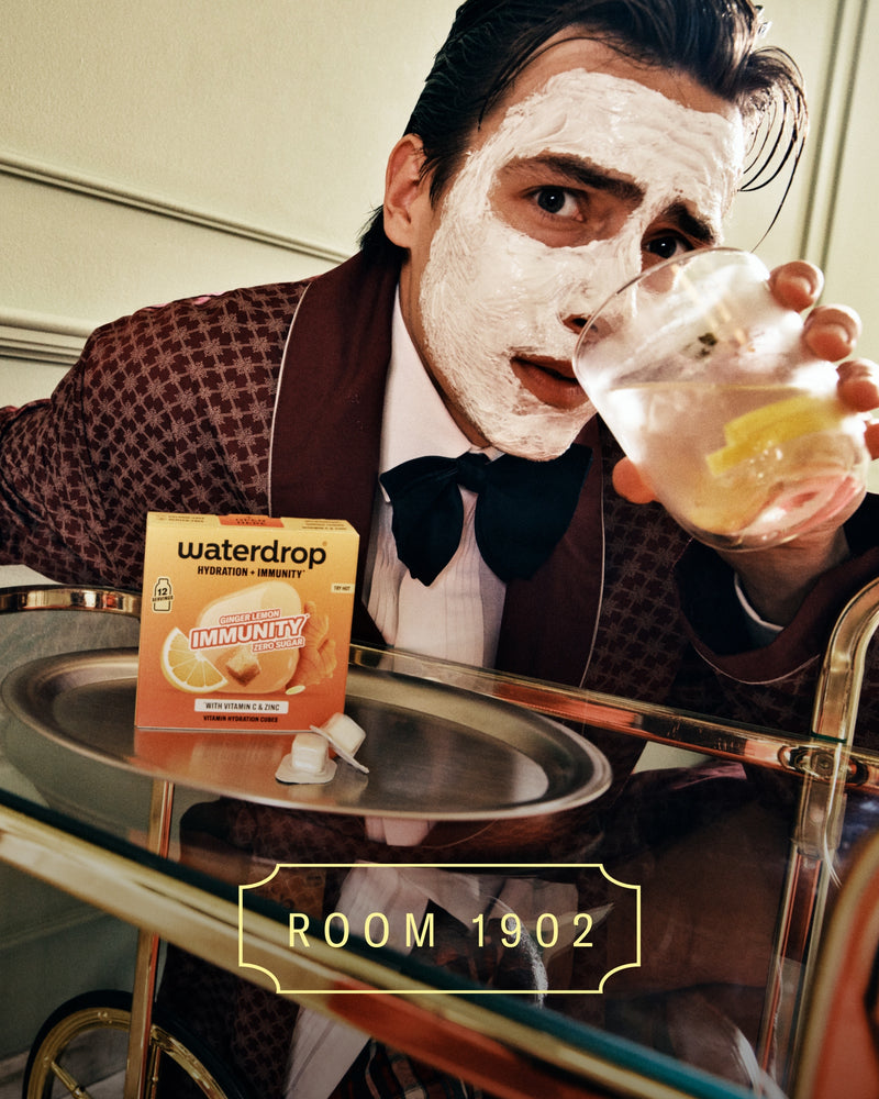 Persona con traje marrón a cuadros y lazo negro, con mascarilla blanca, sostiene un vaso de bebida junto a una bandeja con un producto en caja.