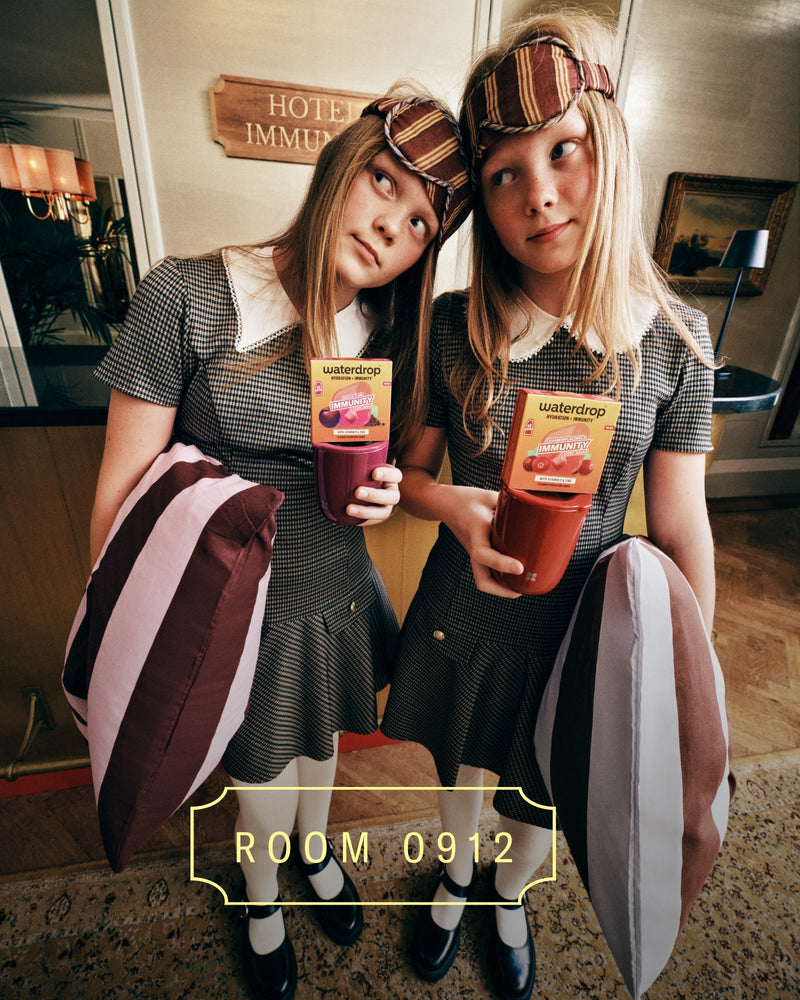 Dos niñas con atuendos a juego y cintas a cuadros sostienen bebidas coloridas en el vestíbulo de un hotel, con bolsas a los lados.