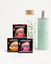 Conjunto de tres cajas Waterdrop flavor boost junto a una botella de vidrio con tapa de madera y un vaso verde claro.