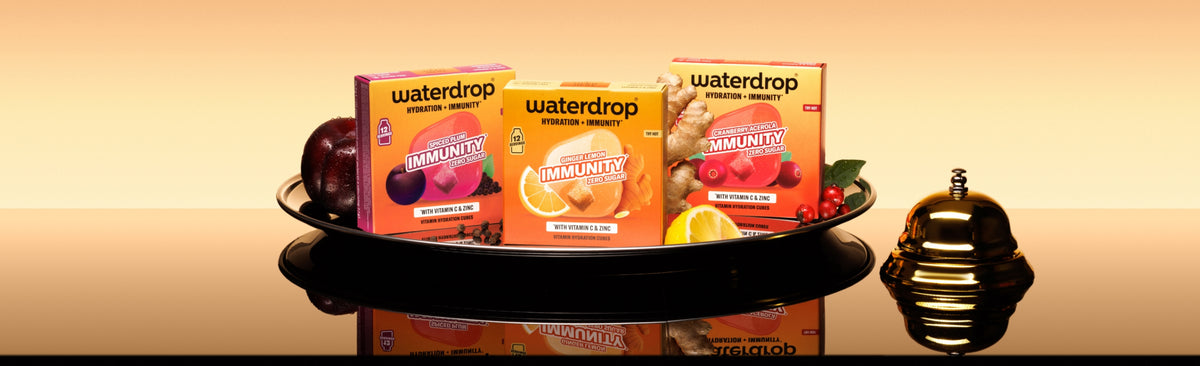 Bandeja con tres cajas Waterdrop sobre una superficie reflectante y una pequeña botella decorativa a la derecha.