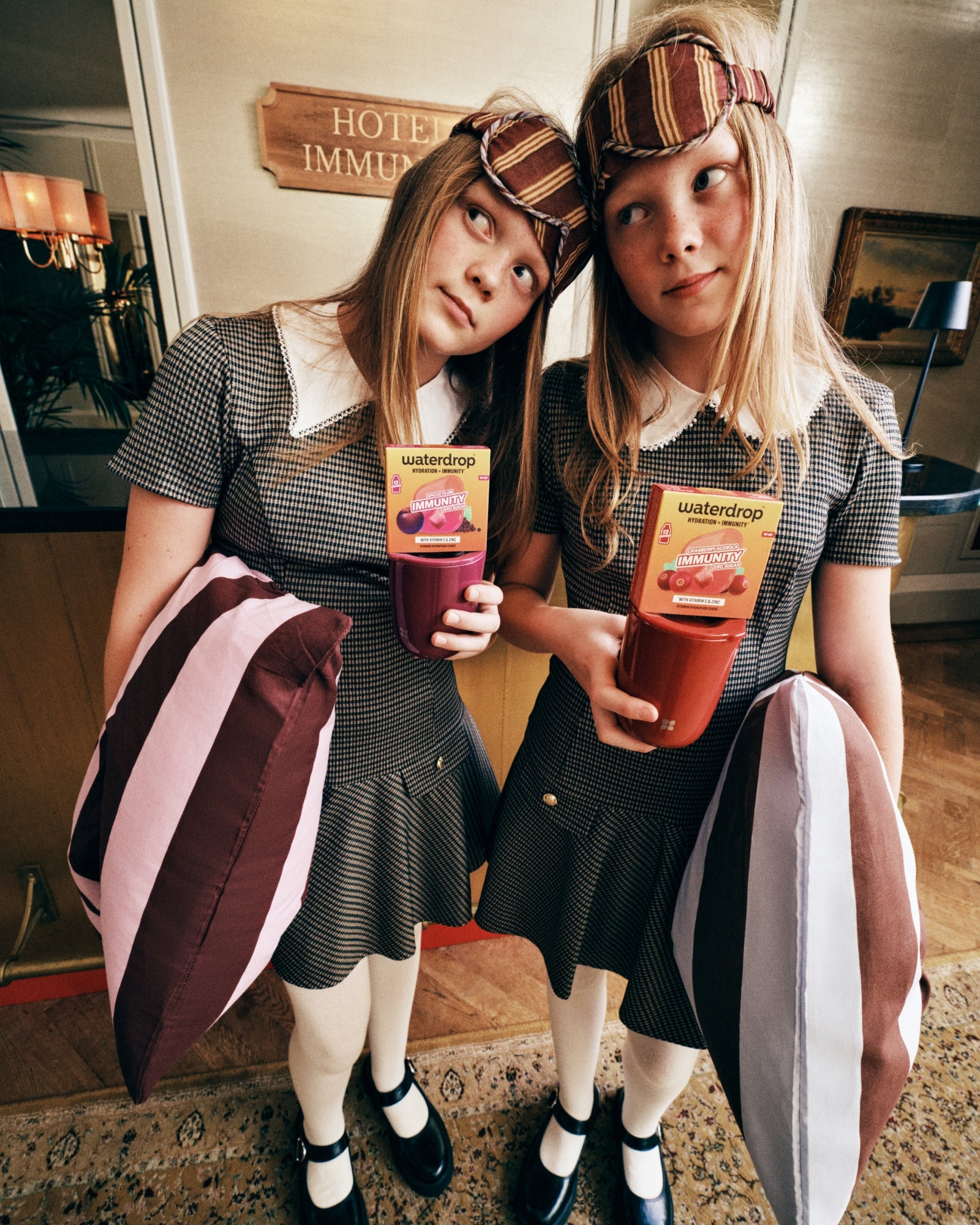 Dos niñas con vestidos grises a juego y cuellos blancos sostienen bebidas de jugo en un vestíbulo de hotel.