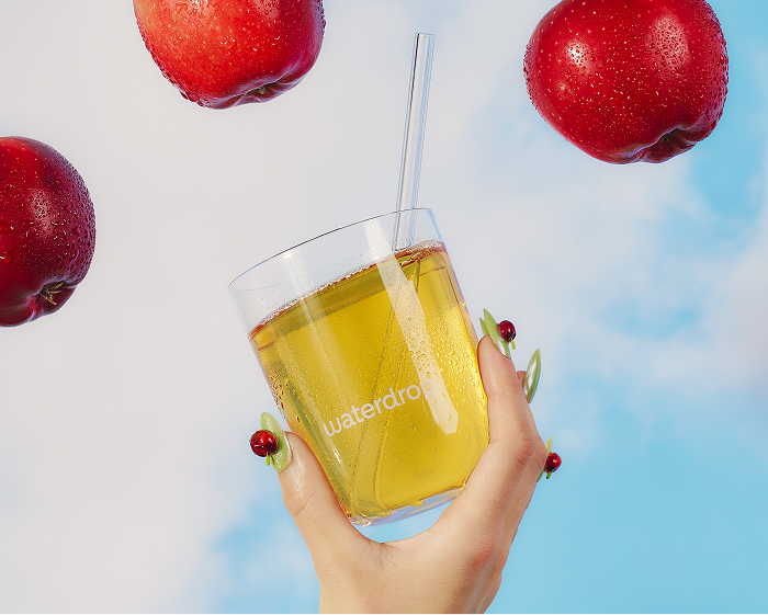 Mano sosteniendo un vaso con jugo amarillo y una pajilla, rodeado de manzanas rojas y un cielo azul.