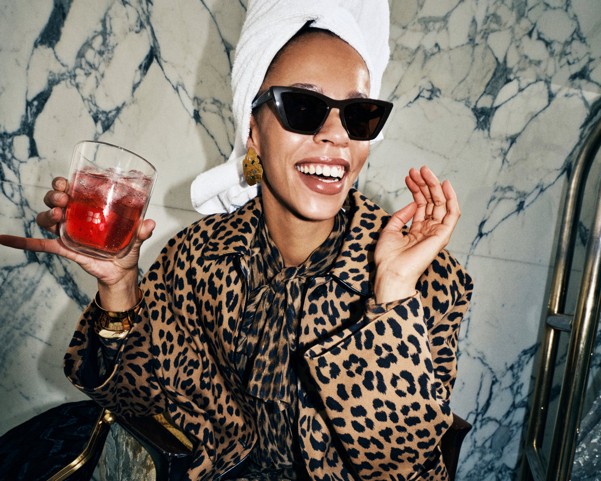 Mujer con abrigo de leopardo y gafas de sol, con una toalla blanca en la cabeza, sosteniendo una bebida roja en un vaso junto a una pared de mármol.