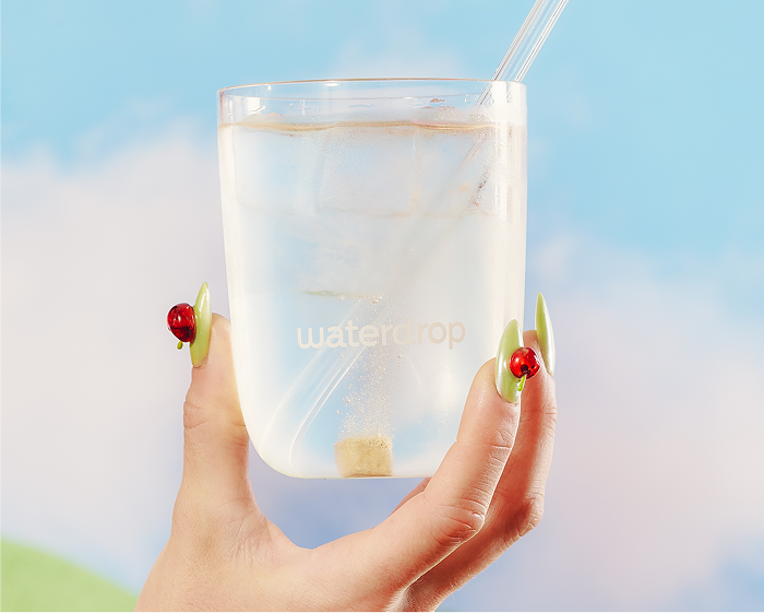 Una mano sostiene un vaso transparente con agua y hielo, con una pajita, frente a un cielo azul.
