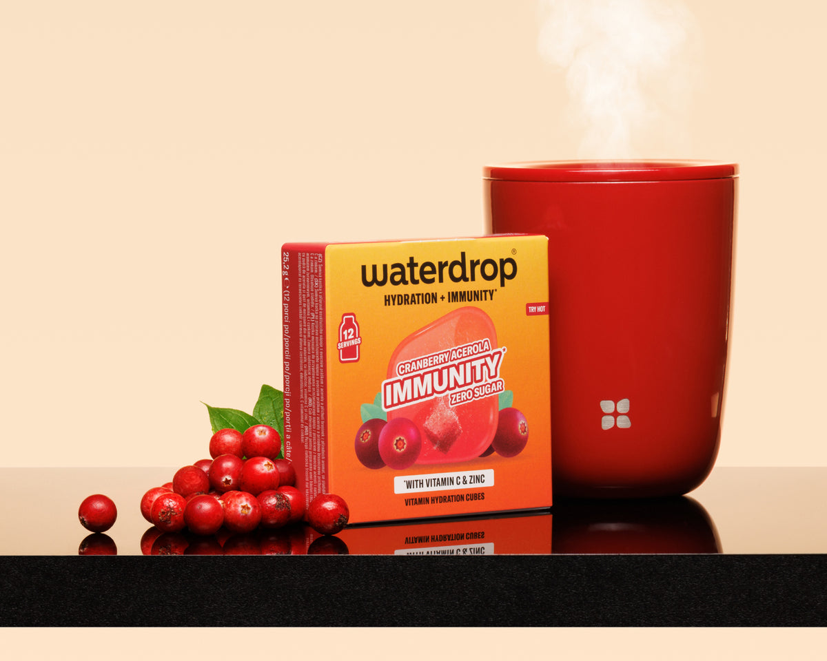 Caja naranja junto a una taza roja humeante y un racimo de bayas rojas sobre una superficie reflectante.