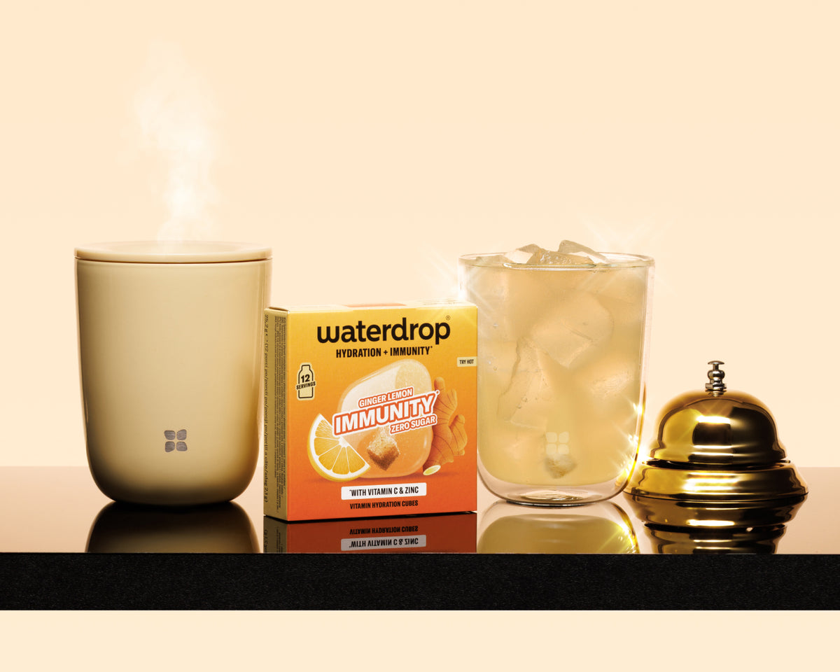 Taza beige humeante, una caja de producto, un vaso con hielo y una campana de bronce sobre una estantería oscura.