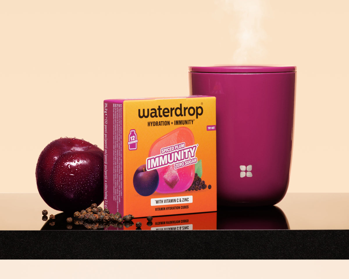 Taza morada humeante junto a una caja naranja Waterdrop, una ciruela y semillas dispersas sobre una superficie reflectante.