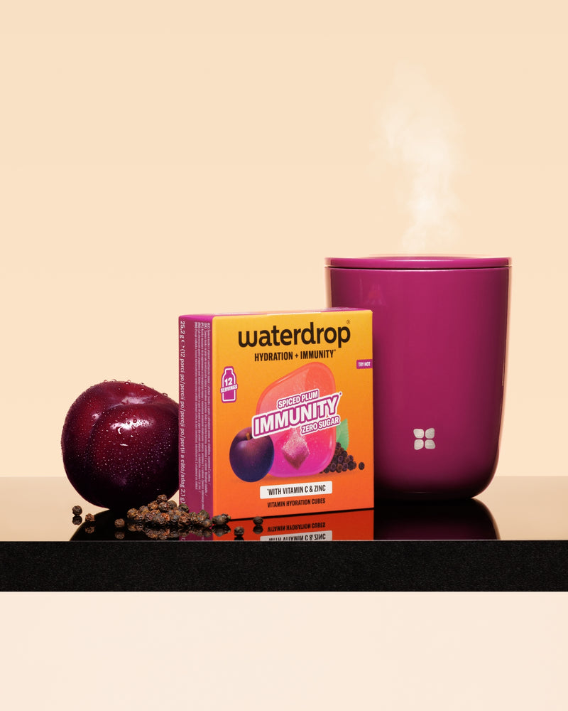 Caja de Waterdrop inmunidad junto a una ciruela y una taza rosa humeante sobre una superficie negra.