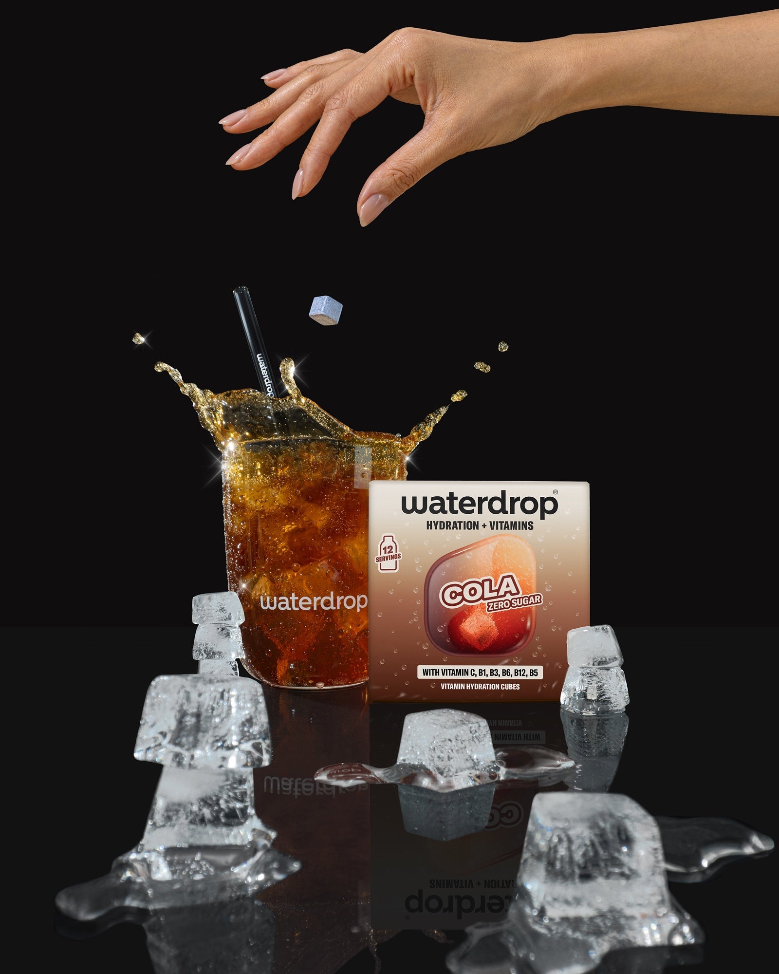Una mano se extiende sobre un vaso de cola con cubitos de hielo, al fondo se ve una caja de bebida.