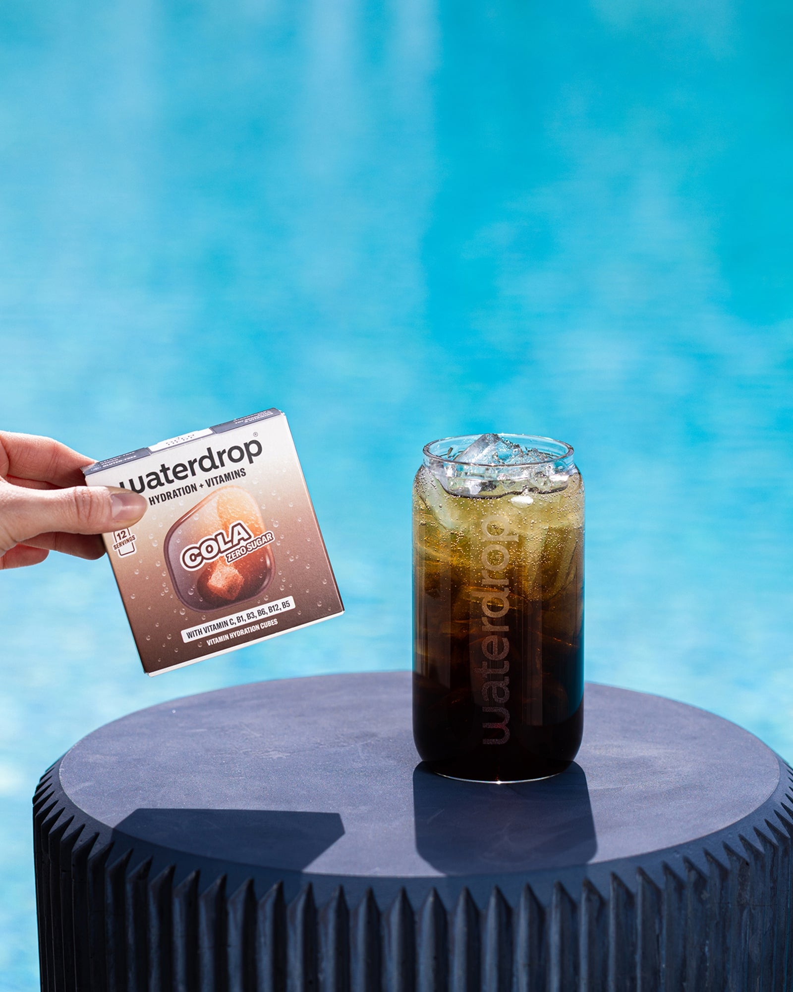 Una mano sostiene un pequeño sobre junto a un vaso con cola fría sobre un pedestal azul circular junto a la piscina.